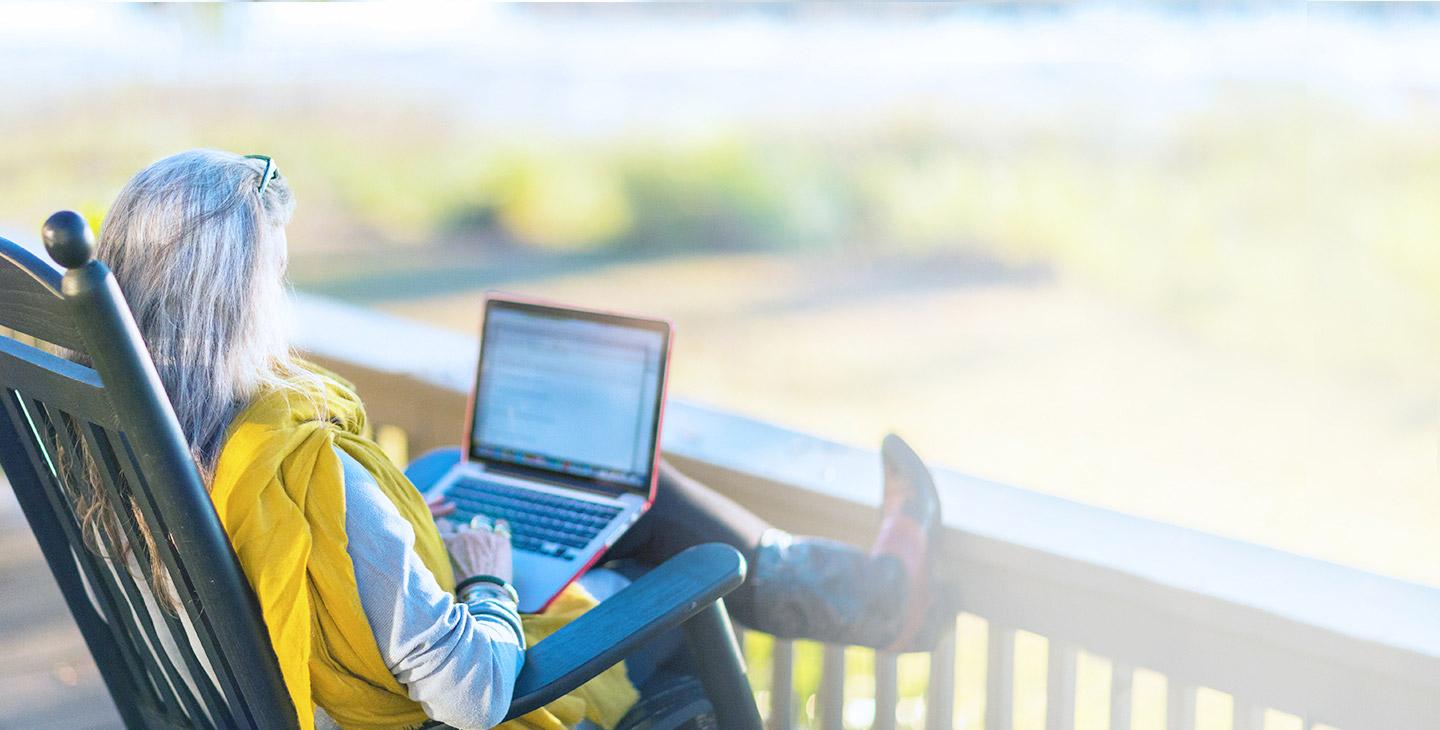 Woman in cowboy boots sitting in a rocking chair on her porch in teh countryside, typing on her laptop